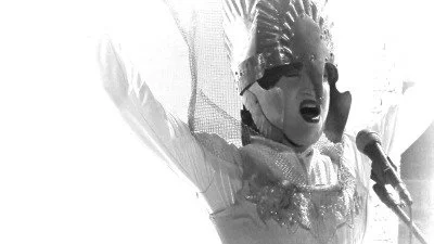 A performer dressed as a clown with a crown, singing passionately into a microphone with arms raised.