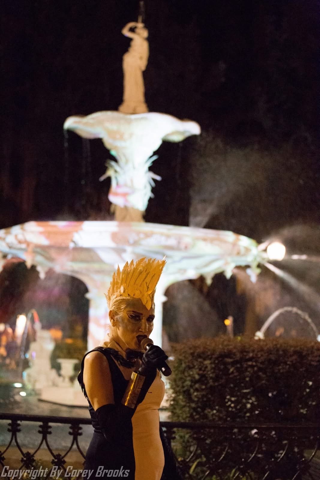 A person dressed in a black and yellow costume with a yellow feathered headpiece stands in front of a lit-up fountain at night, holding a microphone.