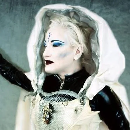 A woman with platinum blonde hair, dramatic makeup with blue eyeshadow and dark lipstick, wearing a white hooded jacket with a metallic chest piece, posed with her eyes closed against a gray background.