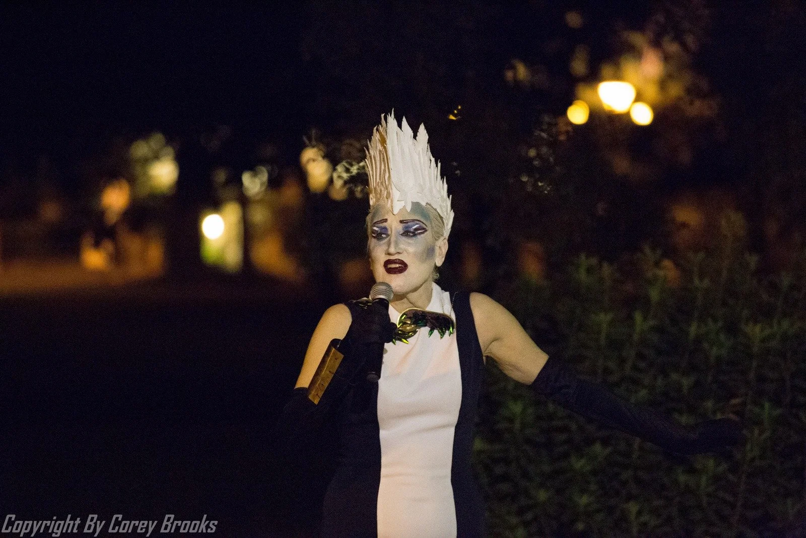 Person in elaborate costume with spiked white headpiece and dramatic makeup holding a microphone, standing outdoors at night.
