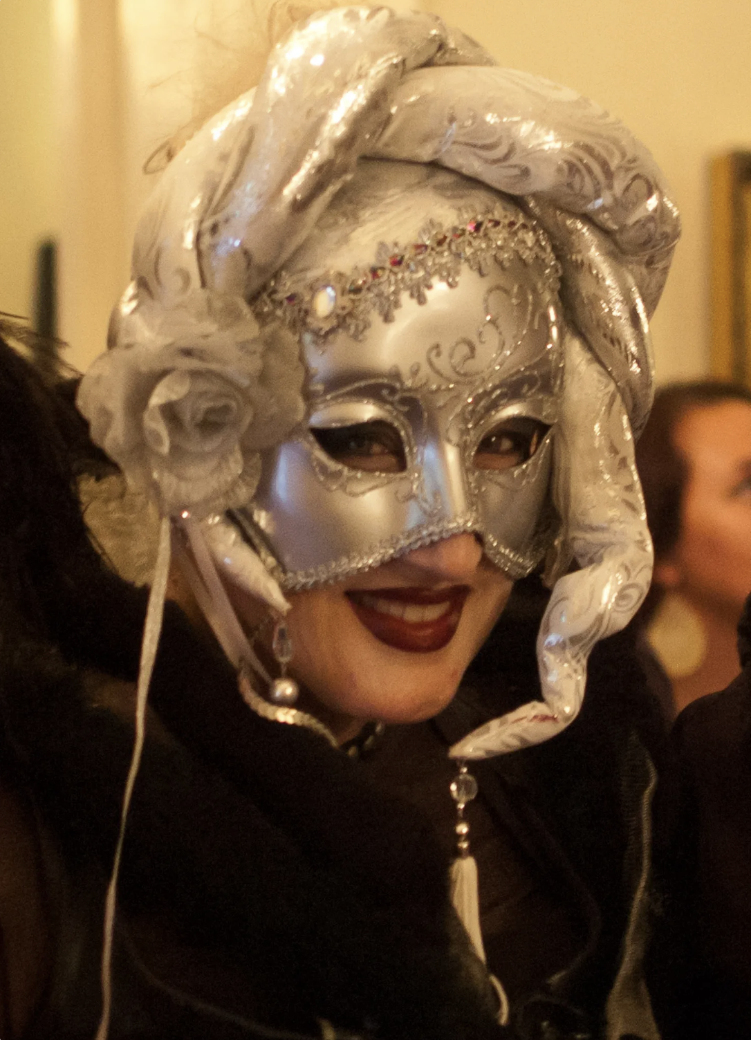 Person wearing a decorative silver Venetian-style mask with intricate designs, a matching head wrap, and dark lipstick, smiling at a celebration or event.