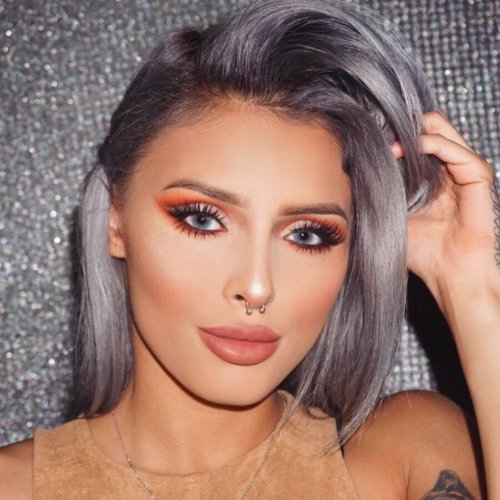 A woman with makeup, gray-dyed hair, and a septum piercing, posing against a glittering silver background.