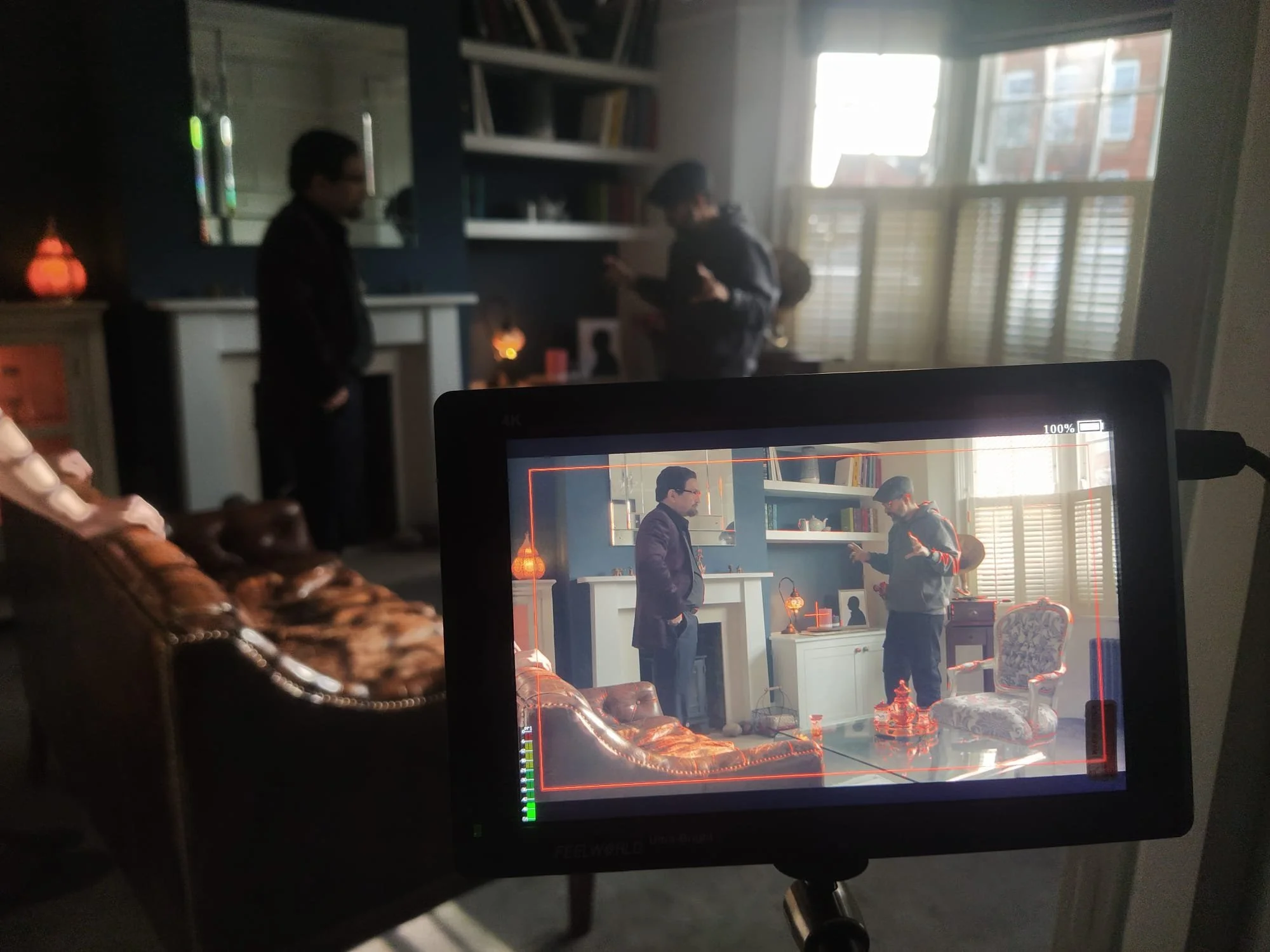 Filming set with two men inside a well-lit living room, with one man on a monitor and the other two in the background.