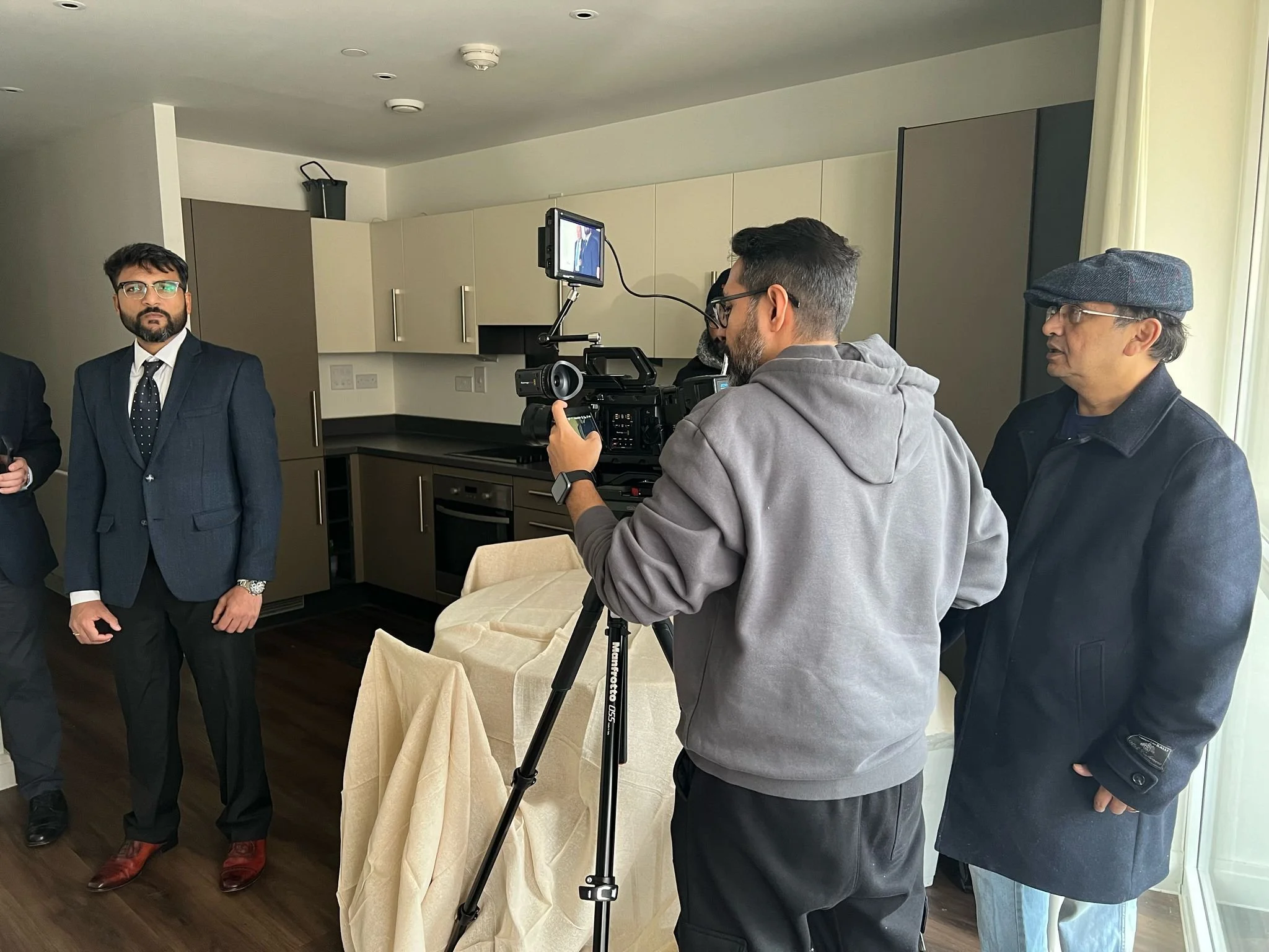 A man in a blue suit and glasses standing in a room being filmed by a cameraman, with another man in a black coat and glasses nearby.