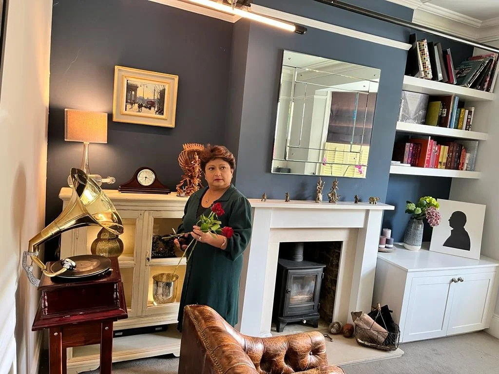 A woman standing in a living room, holding red roses and a phone, beside a vintage record player and a white fireplace with a mirror above it, surrounded by books and decorative items.