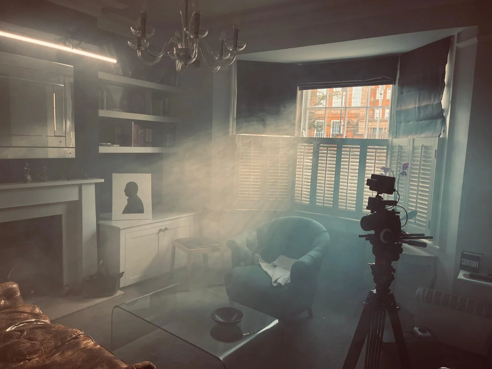 A room filled with natural light from a large window, a camera on a tripod filming the space, a blue armchair with a blanket, a glass coffee table, a brown leather sofa, a fireplace with a silhouette picture above it, and a chandelier hanging from the ceiling, with smoke or fog creating a hazy atmosphere.