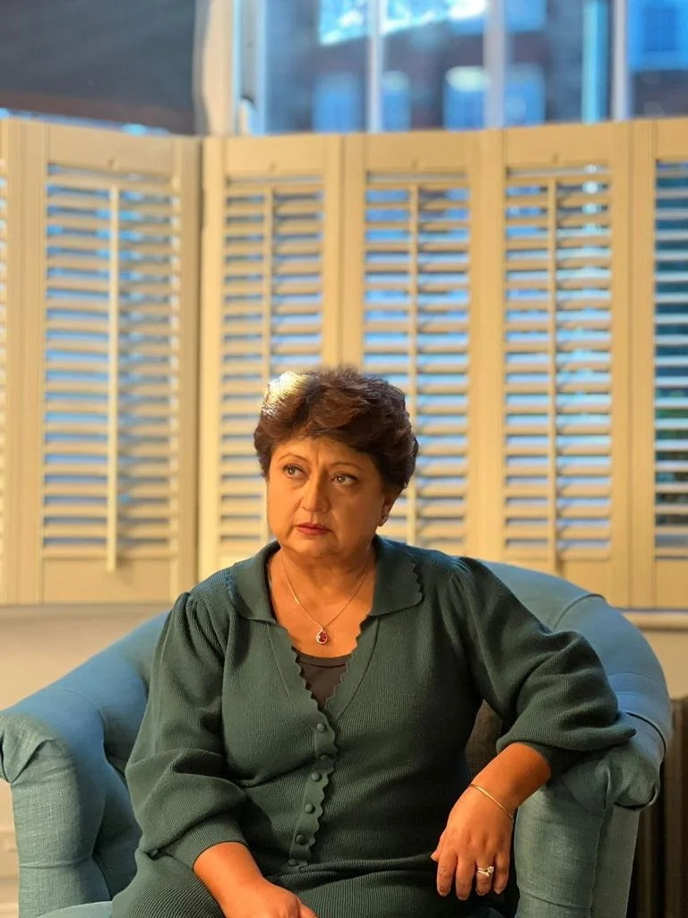 A middle-aged woman with short, dark hair sitting on a blue armchair in front of wooden shutters with a window behind. She is wearing a dark green cardigan with a scalloped edge and a pendant necklace, looking contemplative.
