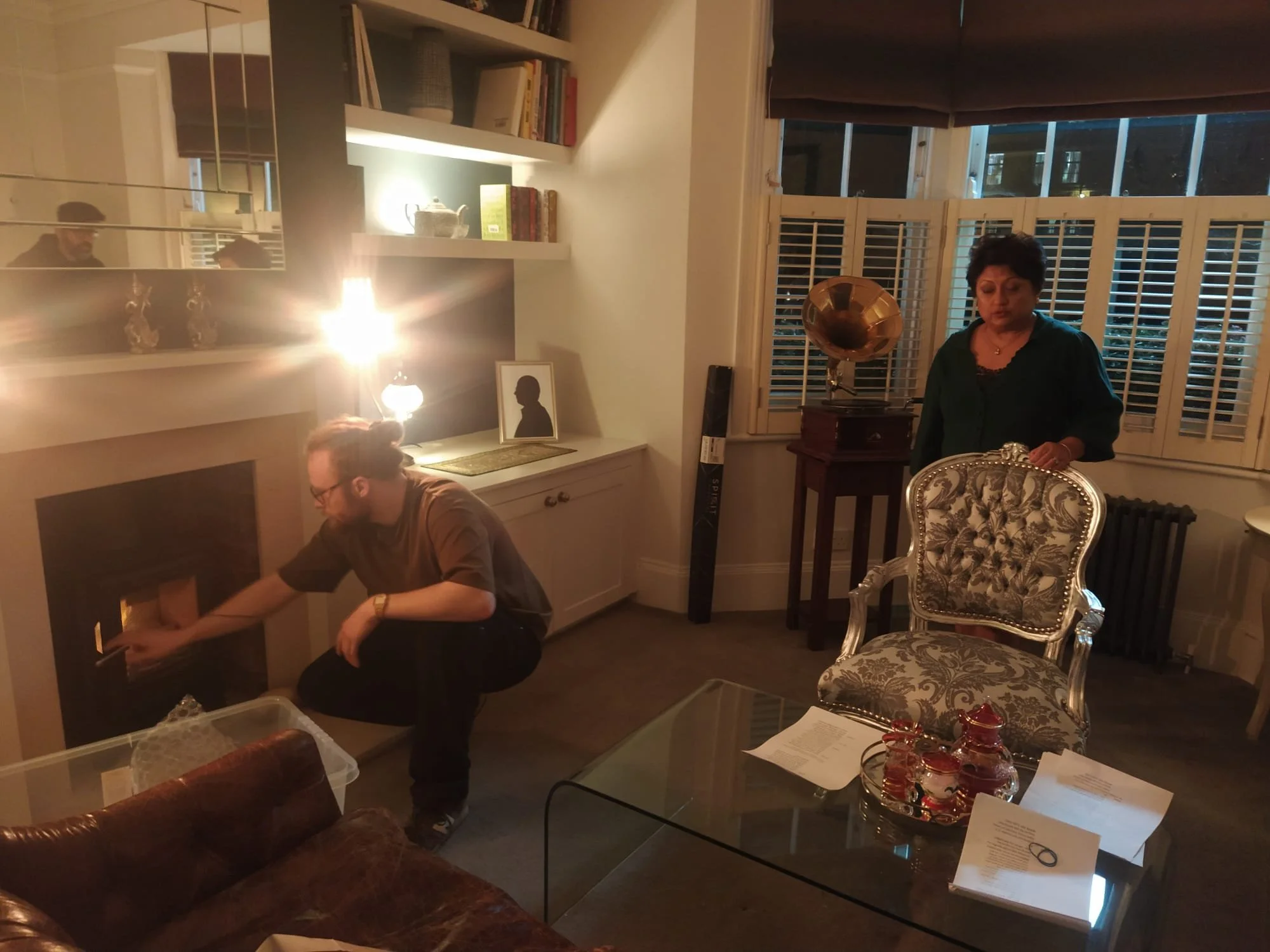 A man kneeling by a fireplace adjusting something, while a woman stands nearby in a cozy, well-decorated living room at night. The room features a patterned armchair, a glass coffee table with papers, a table with red decorative items, a vintage gramophone, and a framed silhouette portrait.