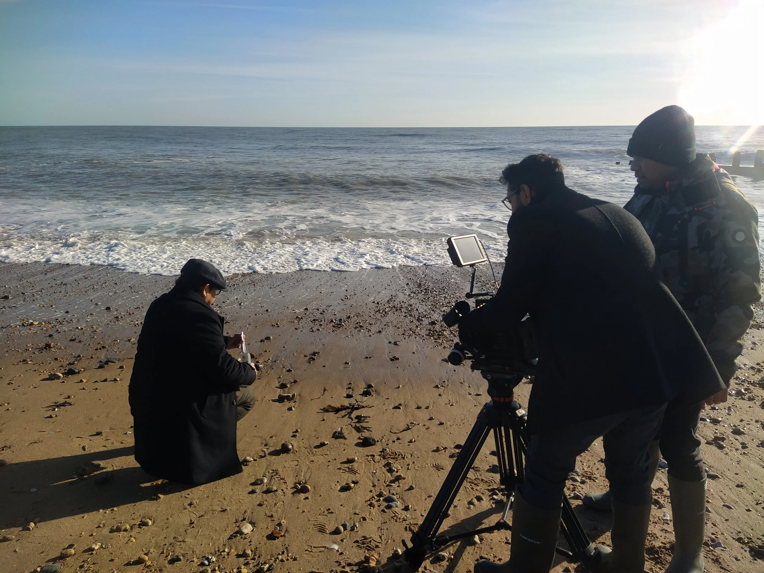 Filmmakers filming a person collecting shells on the beach near the ocean, with crew members operating camera equipment.