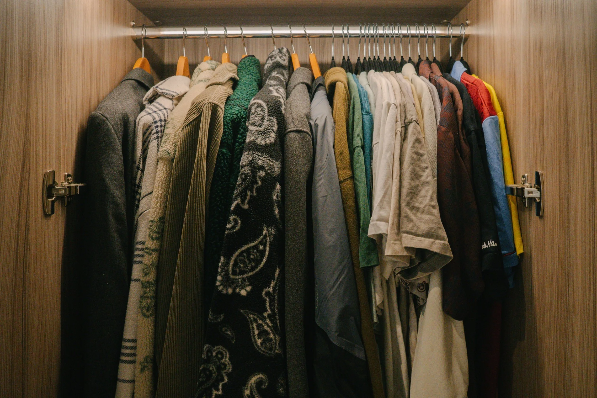 A wardrobe with various jackets and coats hanging on a metal rod, organised by colour from dark on the left to light and colourful on the right.