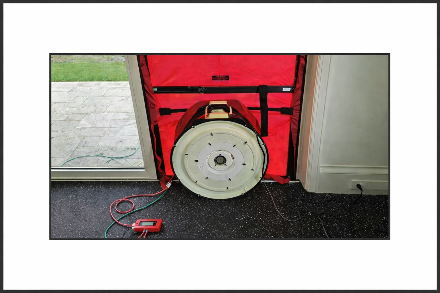 A large circular device used for cleaning ducts is placed on a black speckled floor inside a room near a sliding glass door, connected to a red and black electrical device with green and red cables.