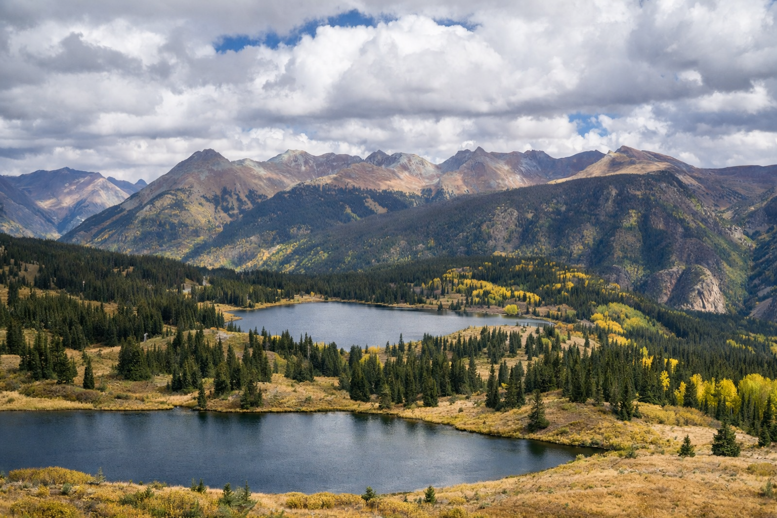 Scenic landscape of two lakes in a valley surrounded by forested hills and mountains under a cloudy sky.