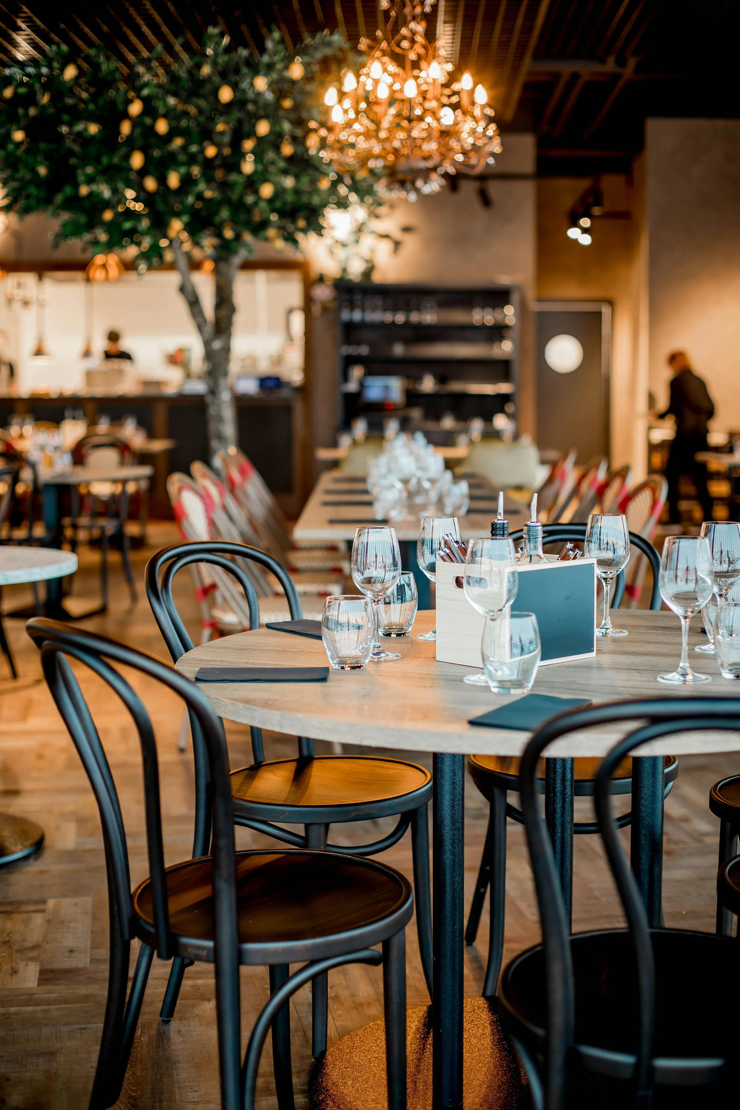 Intérieur d'un restaurant élégant avec des tables dressées, des chaises en bois, un lustre brillant et un arbre décoratif.
