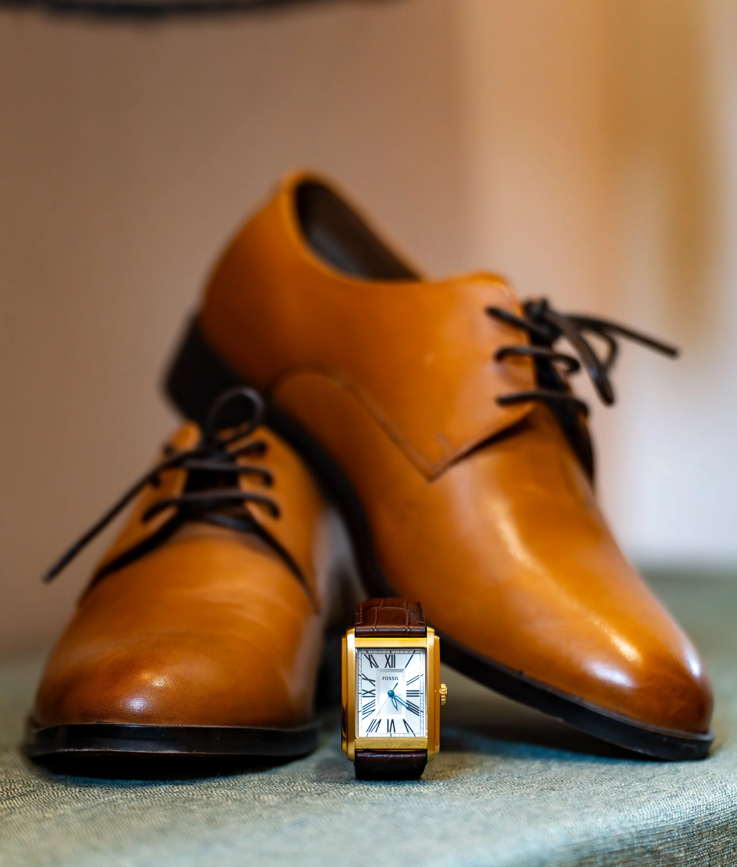 Chaussures en cuir marron foncé et montre-fossil à cadran rectangulaire avec chiffres romains, posés sur un tissu gris.