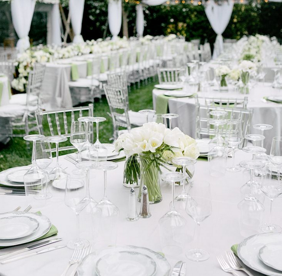 Tables élégamment dressées pour un événement en plein air, avec des chaises transparentes, nappes blanches, sets de table verts, verres à pied et bouquet de fleurs blanches au centre. Ambiance festive et sophistiquée.