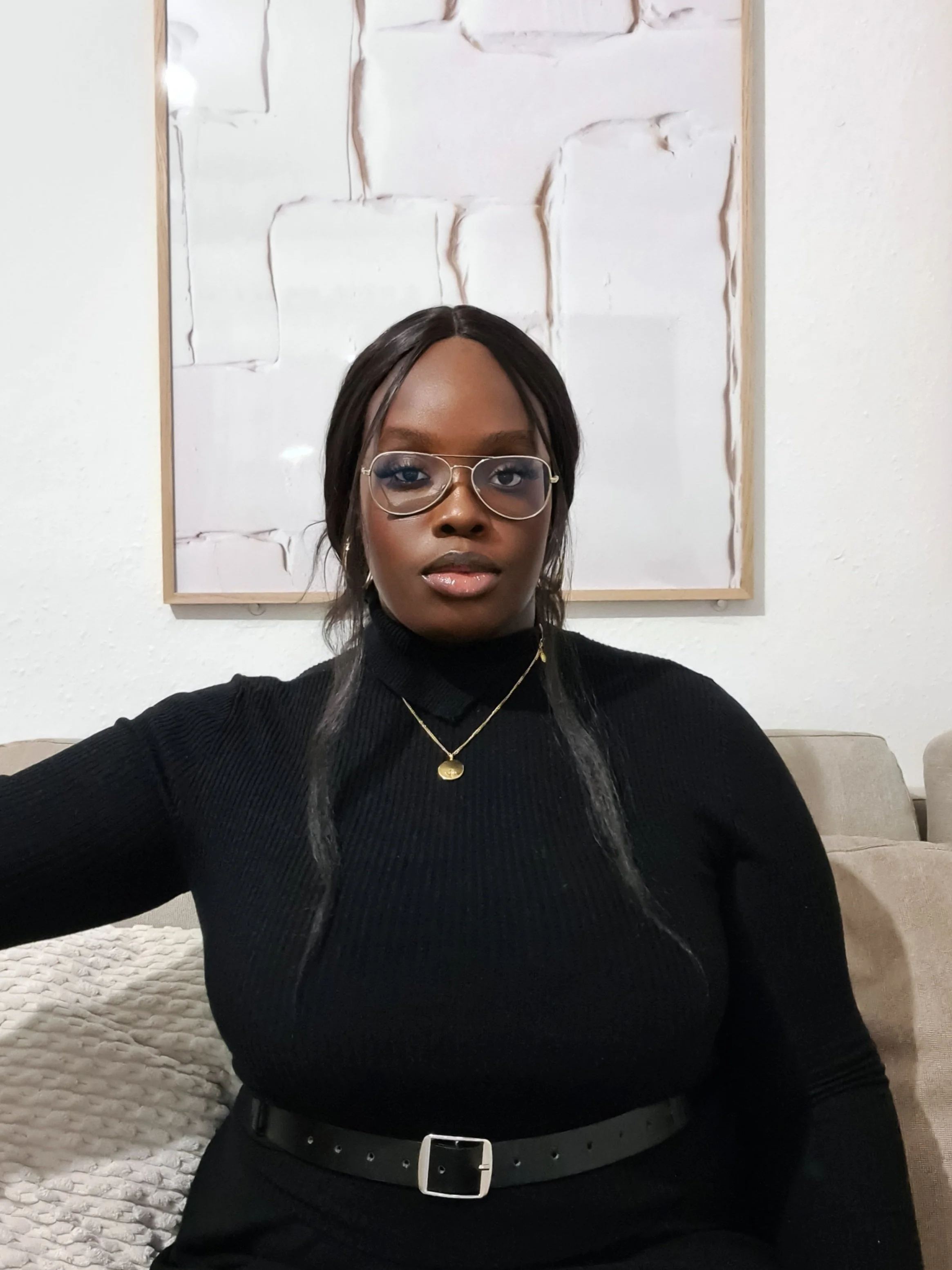 Une personne portant des lunettes et un pull noir est assise sur un canapé devant un tableau blanc. Elle porte un collier doré et une ceinture noire.