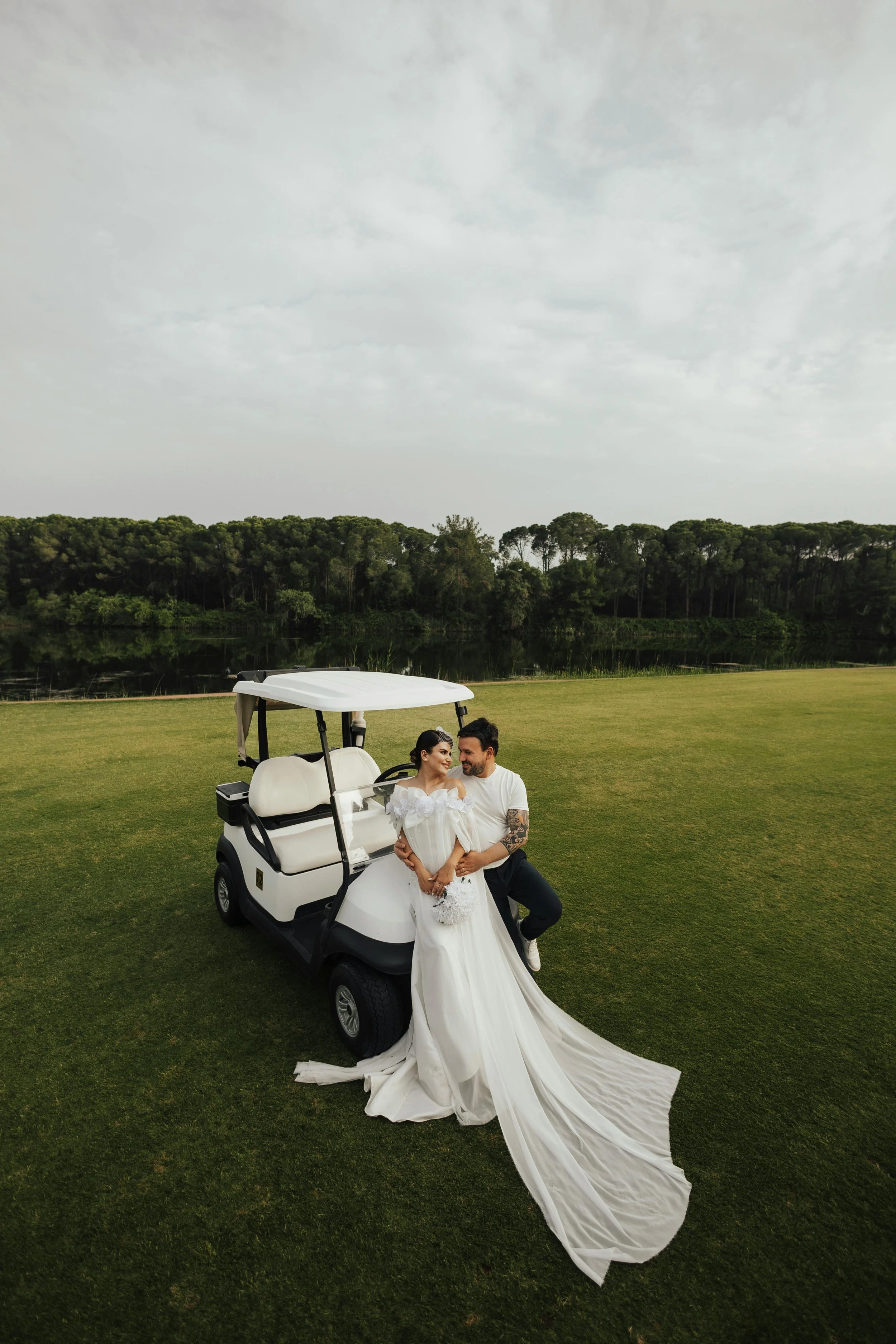 Un couple posé près d'une voiturette de golf sur une pelouse verte, avec des arbres en arrière-plan. La femme porte une robe de mariée.