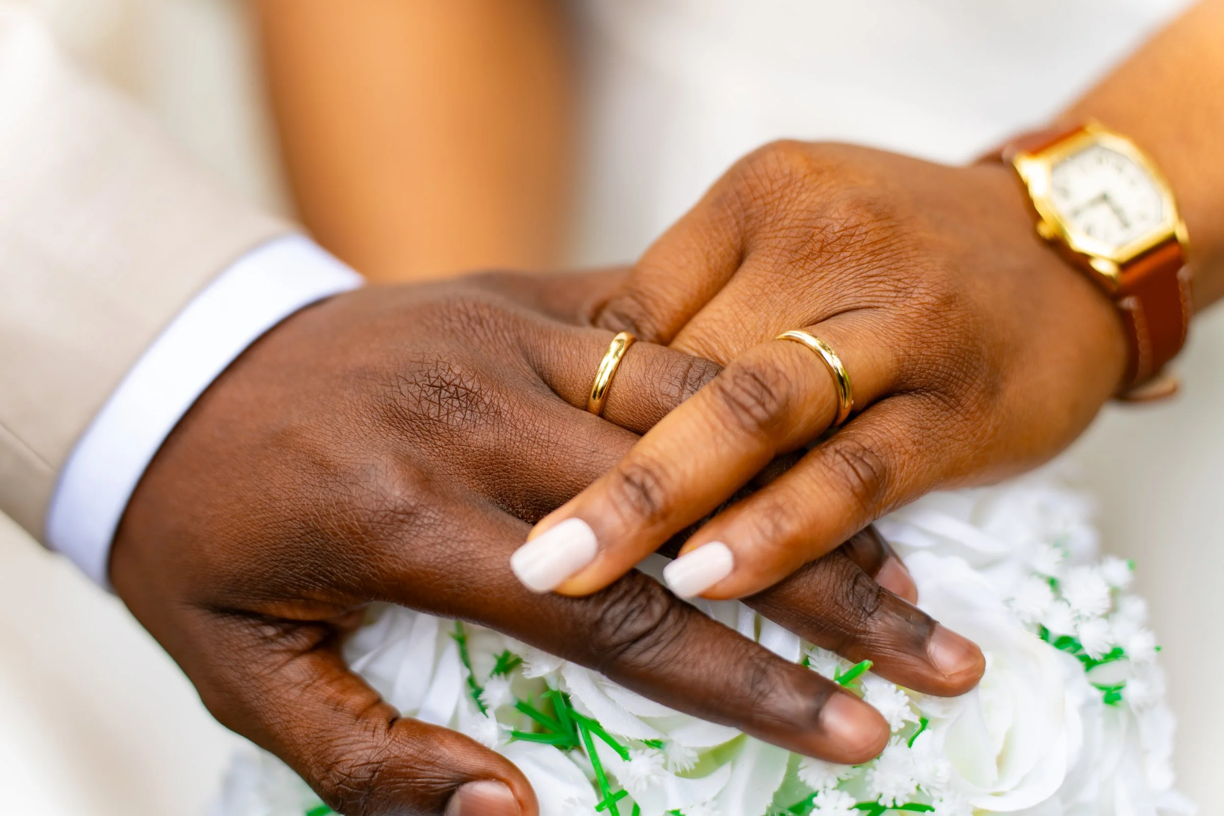 Deux mains de personnes différentes, dont une porte une montre en or et une bague en or, se tiennent l'une l'autre, au-dessus d'une décoration florale.