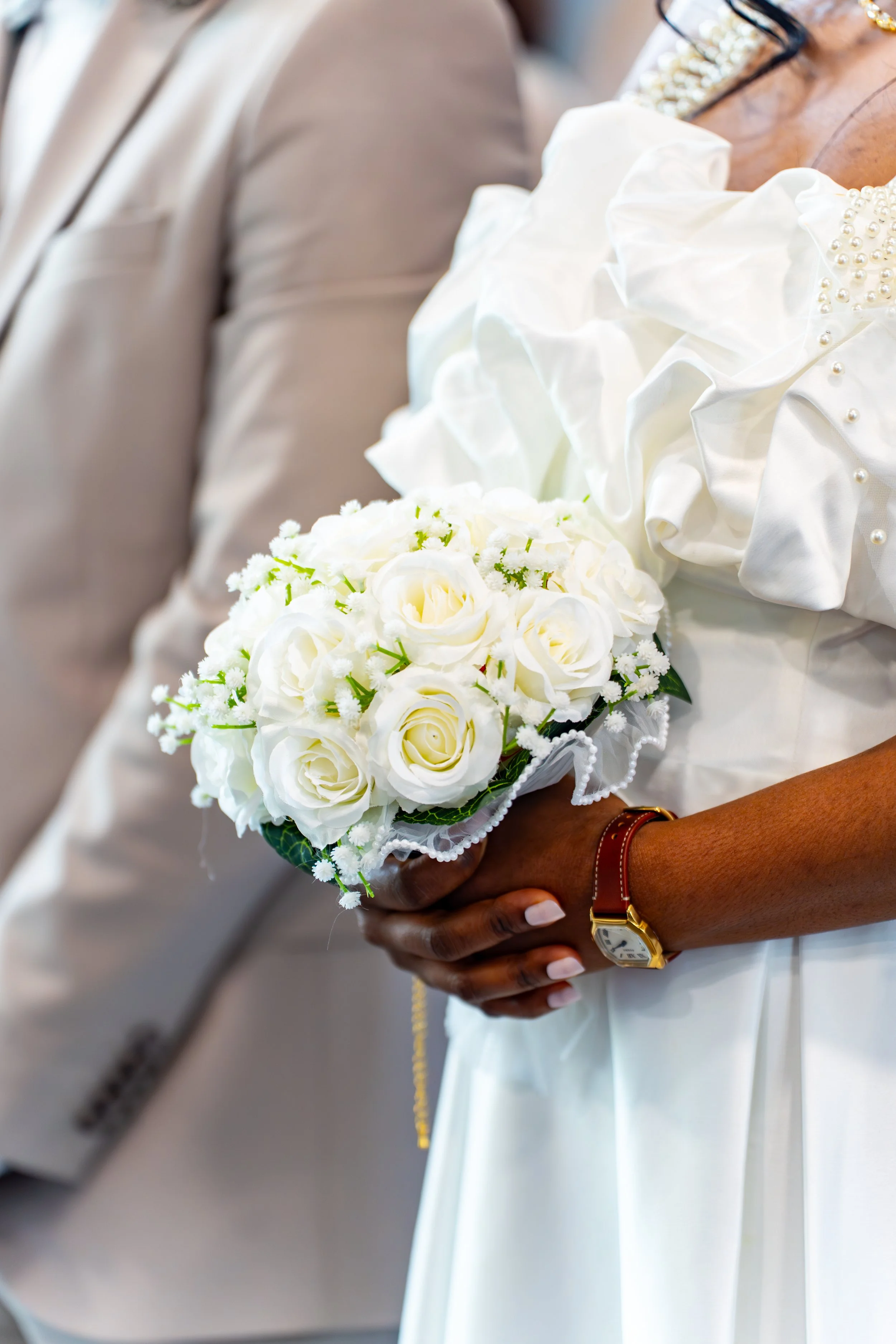 Femme tenant un bouquet de fleurs blanches à son mariage à côté de son mari. Robe blanche avec détails en perles, montre en or avec bracelet en cuir, fond flou.