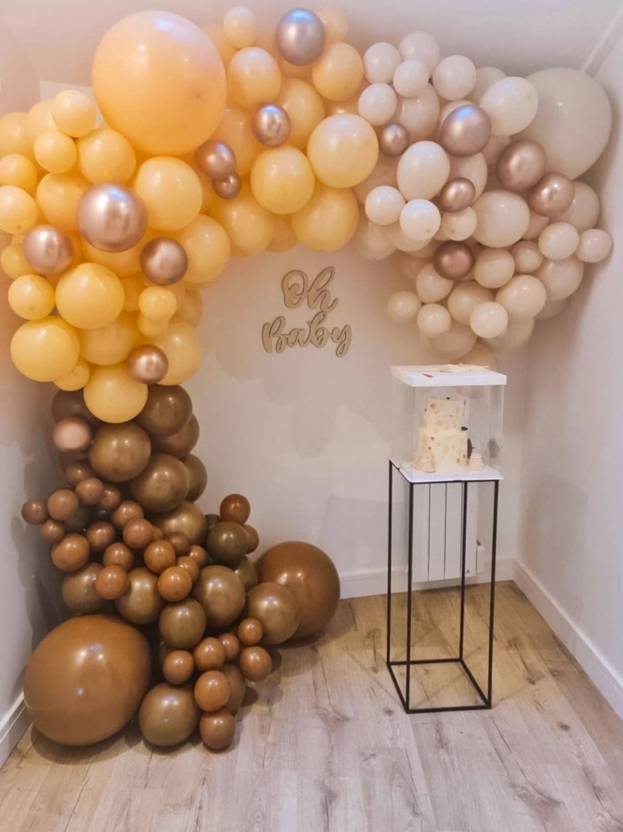Décoration composée de ballons de couleur crème, jaune et dorée formant une arche. Il y a une inscription "Oh baby" en arrière-plan et une table avec un gâteau sur la droite. Le sol est en bois clair et les murs sont blancs baby shower, gender reveal
