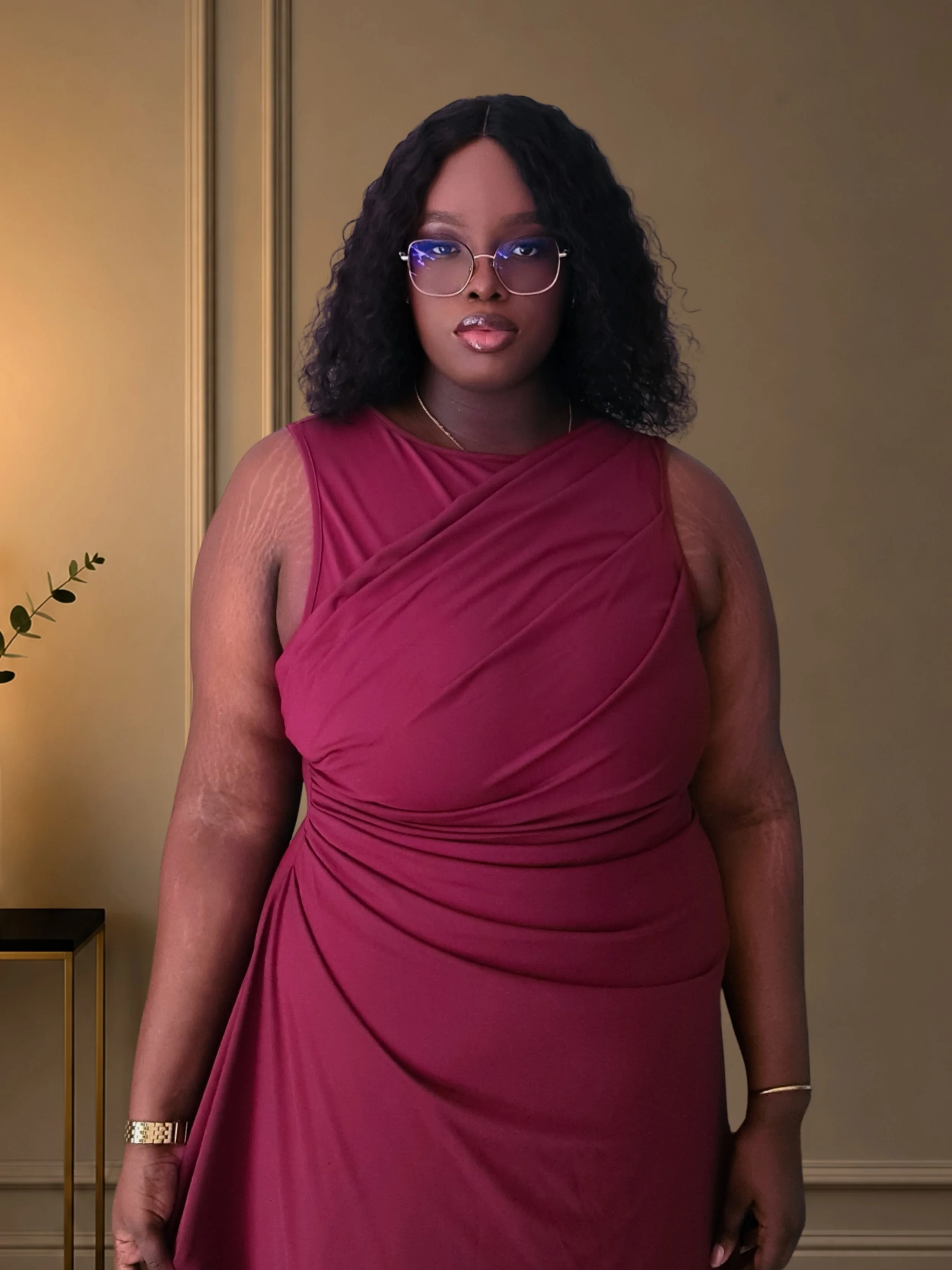 Une femme portant une robe rouge foncé, avec des lunettes rondes, se tenant dans une pièce aux murs beige avec moulures ornementales.