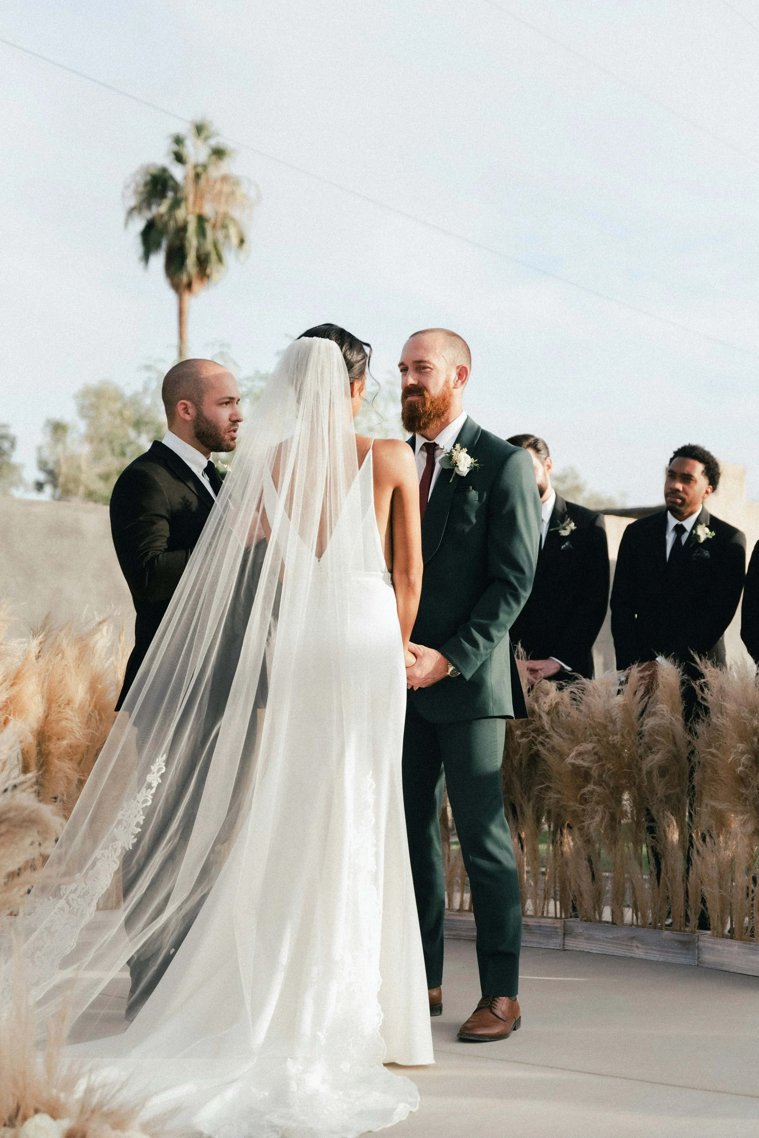 Cérémonie de mariage en plein air avec un couple échangeant des vœux devant un officiant, entouré de spectateurs en costume et une végétation sèche en arrière-plan.