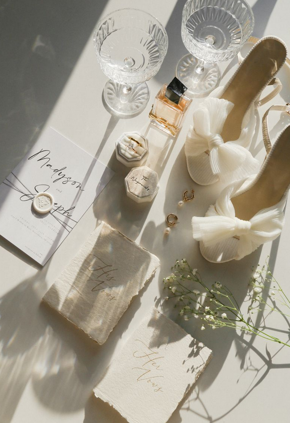 Accessoires de mariage comprenant deux verres à pied, une bouteille de parfum, deux boîtes à bijoux, des bijoux, des chaussures blanches avec nœuds, fleurs blanches, et cartes de vœux de mariage."