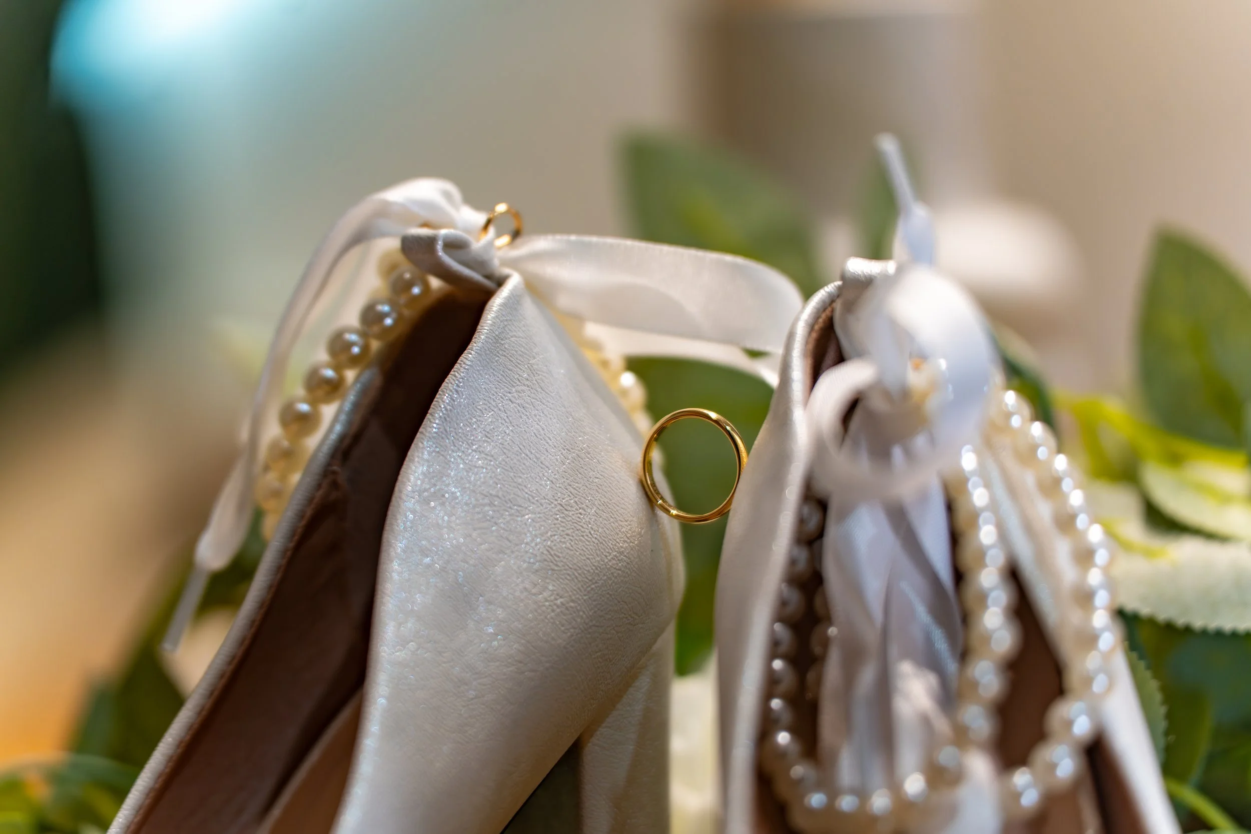 Une paire de chaussure avec des détails de perles, présentée de face avec un focus sur une bague de mariage dorée accrochée.
