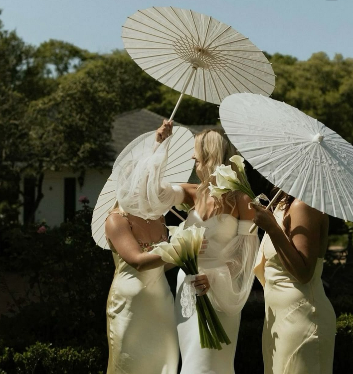 Trois femmes en robes de mariée crème tenant des parasols et des bouquets de fleurs blanches dans un jardin verdoyant.