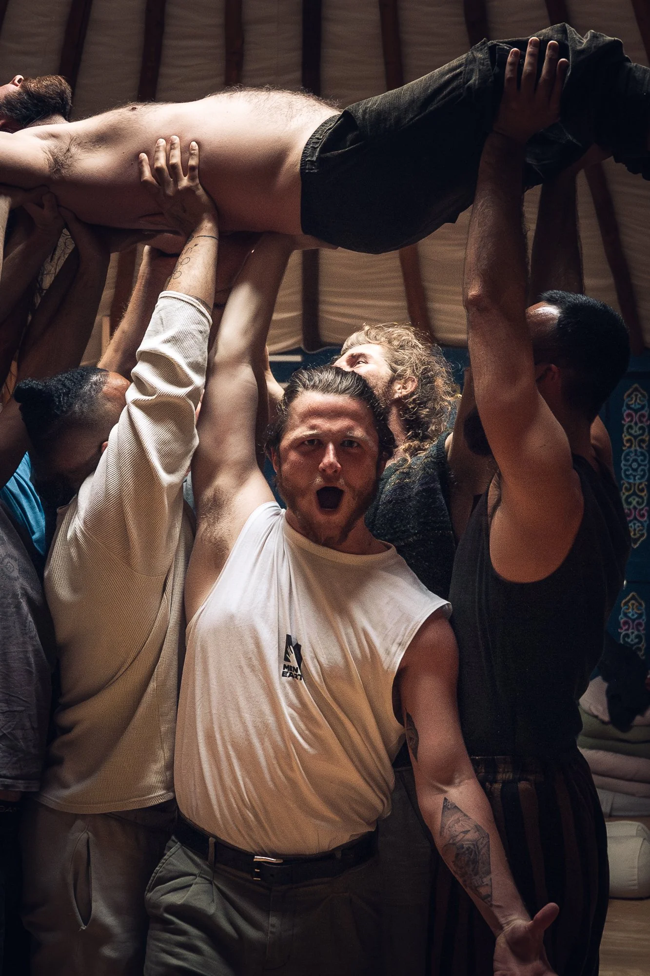Group of people supporting a shirtless man lifted above their heads in a celebration or ritual, inside a wooden structure.