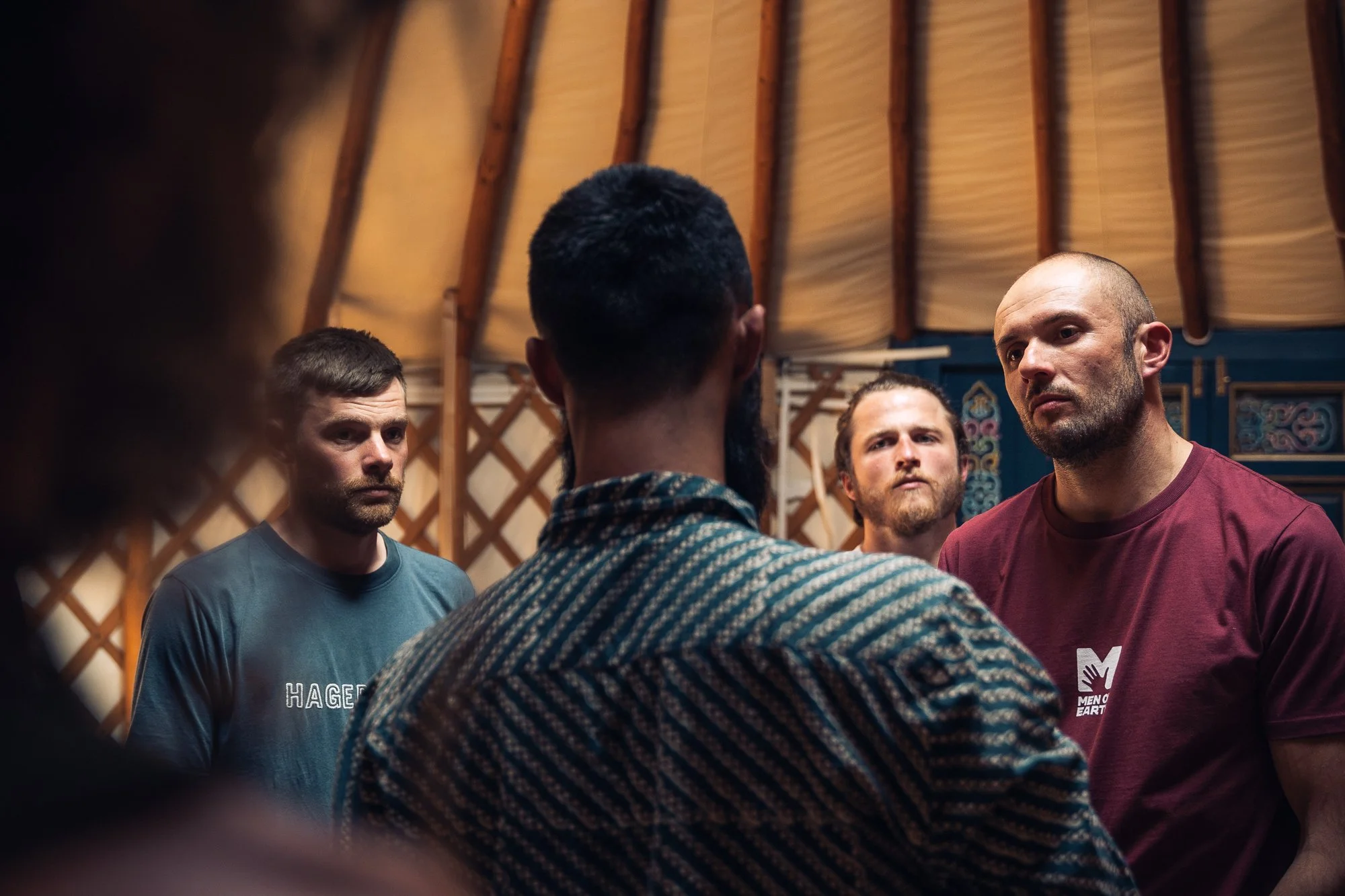 Four men are standing in a circle, engaged in what appears to be a serious discussion or conversation inside a yurt with wooden lattice walls and a beige fabric ceiling.