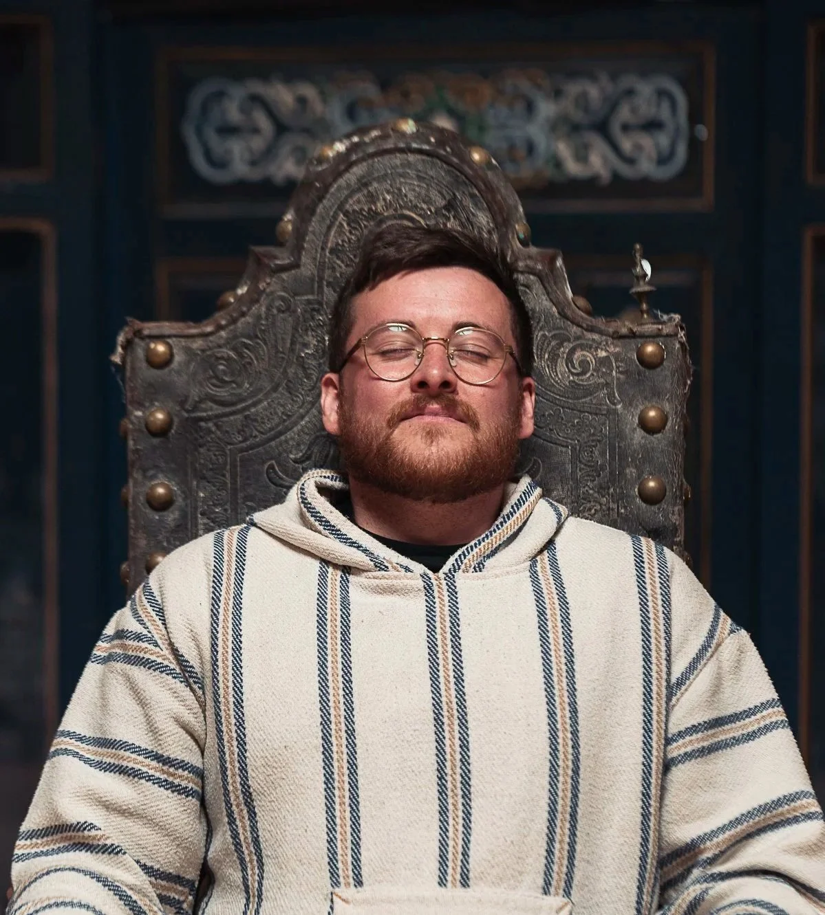 A man with glasses and a beard sits on a throne, wearing a striped hoodie, with eyes closed and a calm expression, in a dimly lit room with ornate decor.