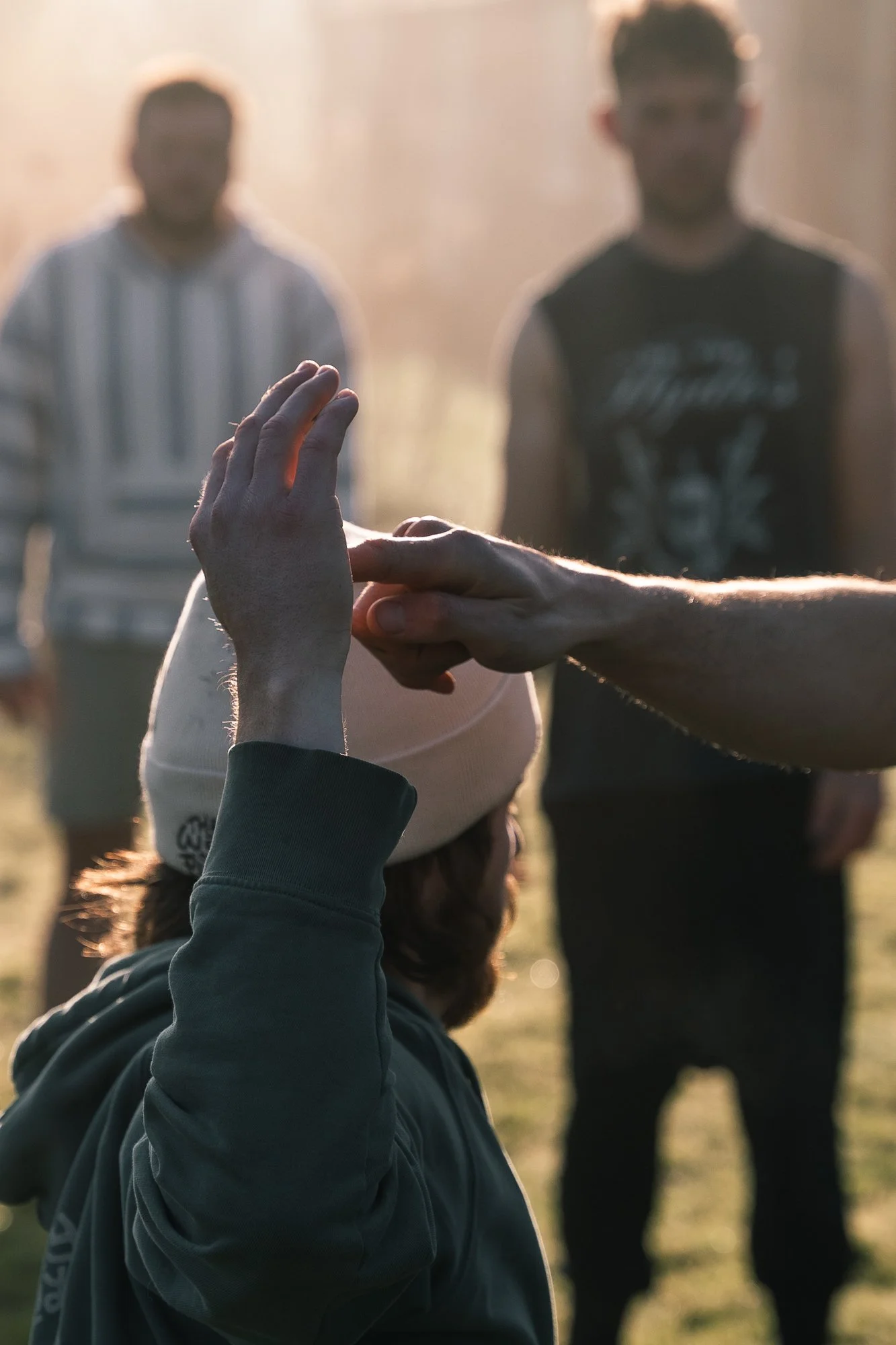 Two people in the background standing outdoors as a third person in the foreground, wearing a beige beanie and a hoodie, is receiving a handshake or high five, with sunlight behind them.