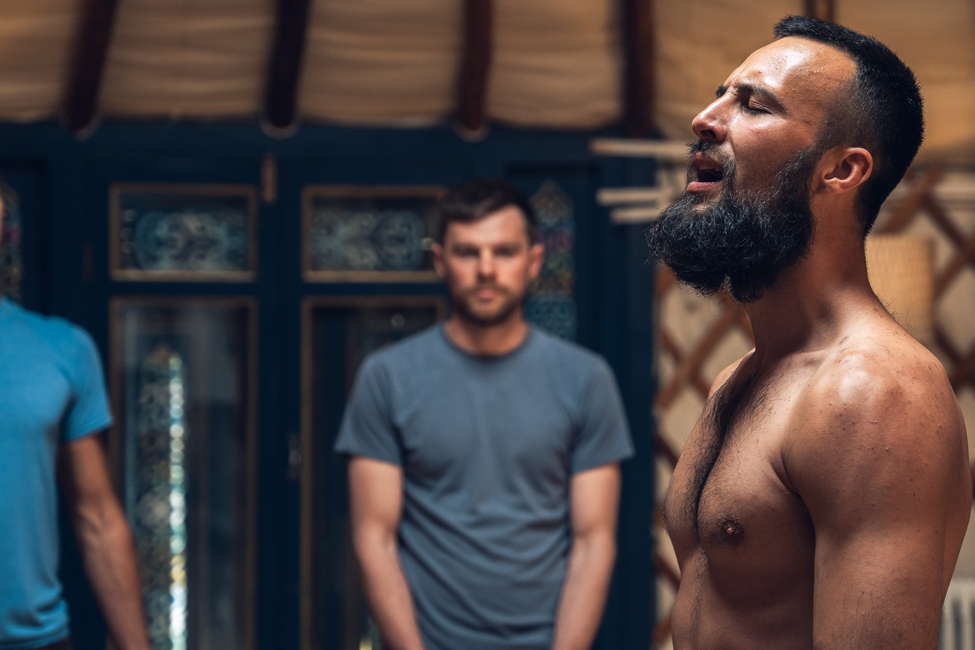 A shirtless man with a beard singing passionately in a room with two other men, one in a blue shirt and the other in a gray shirt, in the background.