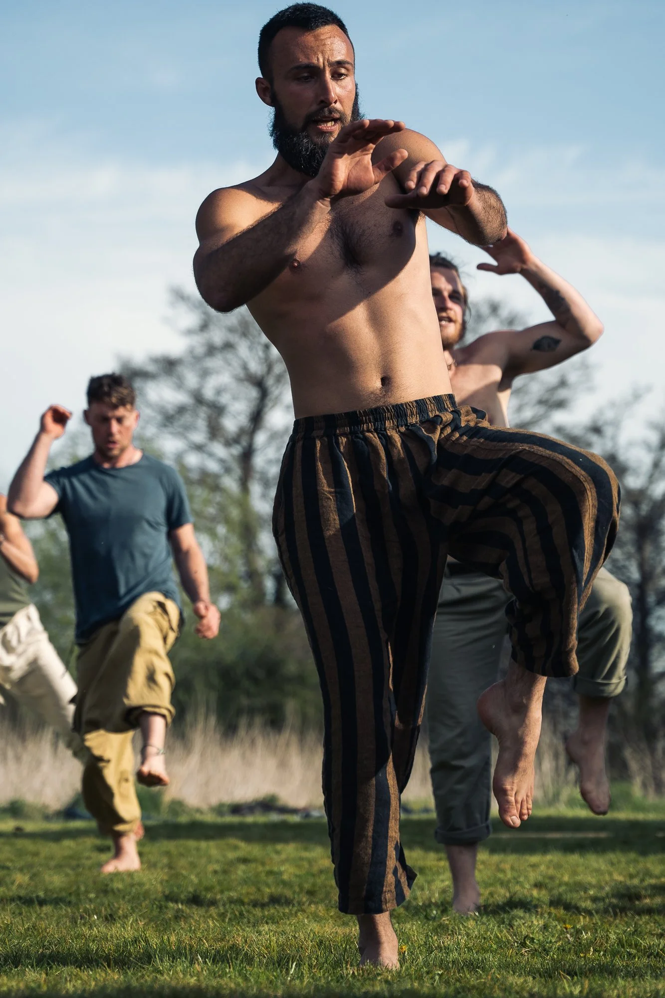 A group of men practicing martial arts outdoors on grass, with one man in the foreground raising his knees and arms, shirtless and wearing striped pants.