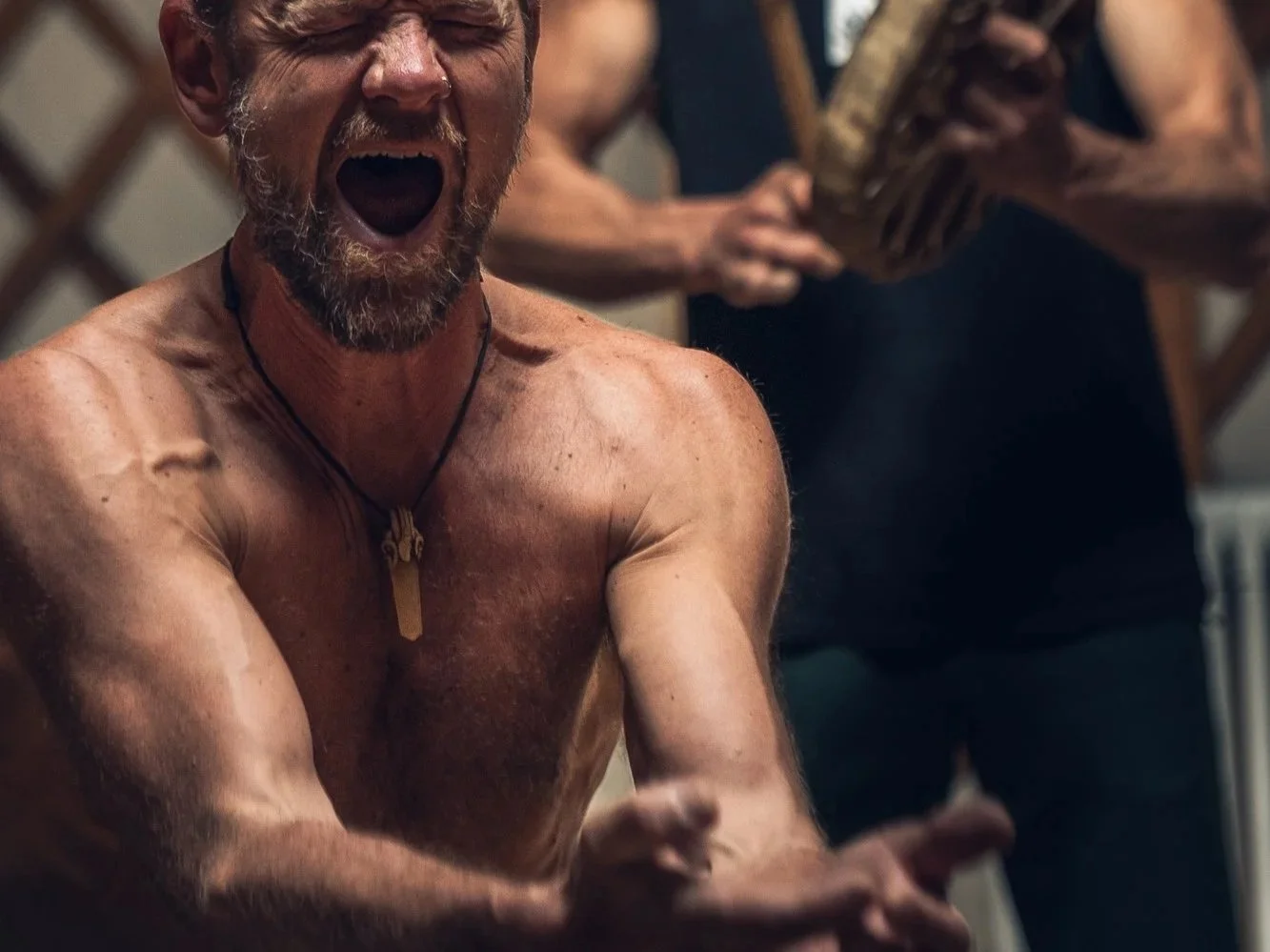 A shirtless middle-aged man with a beard and short hair, wearing a necklace, appears to be shouting or cheering enthusiastically, with his arms extended and fists clenched. In the background, a person is holding a wooden object.