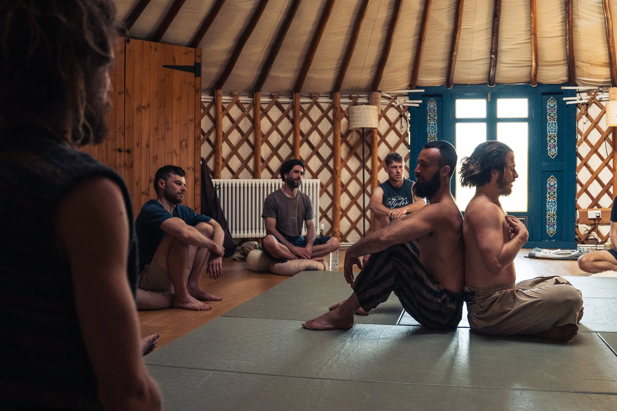 Group of people sitting on the floor in a circle inside a rustic wooden room, possibly practicing meditation or yoga.