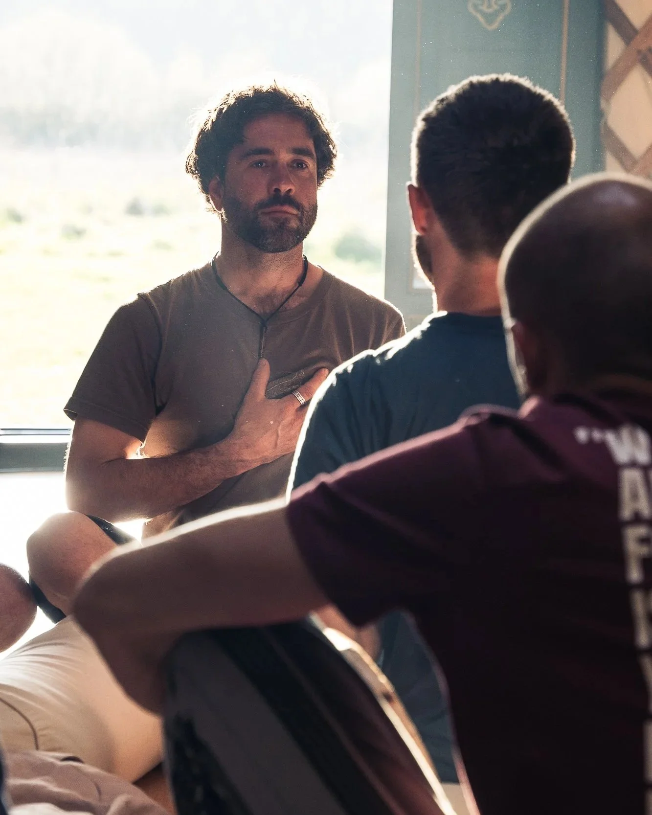 A man with a beard and brown hair holding his hand over his heart, standing in front of a window, speaking to a group of people.