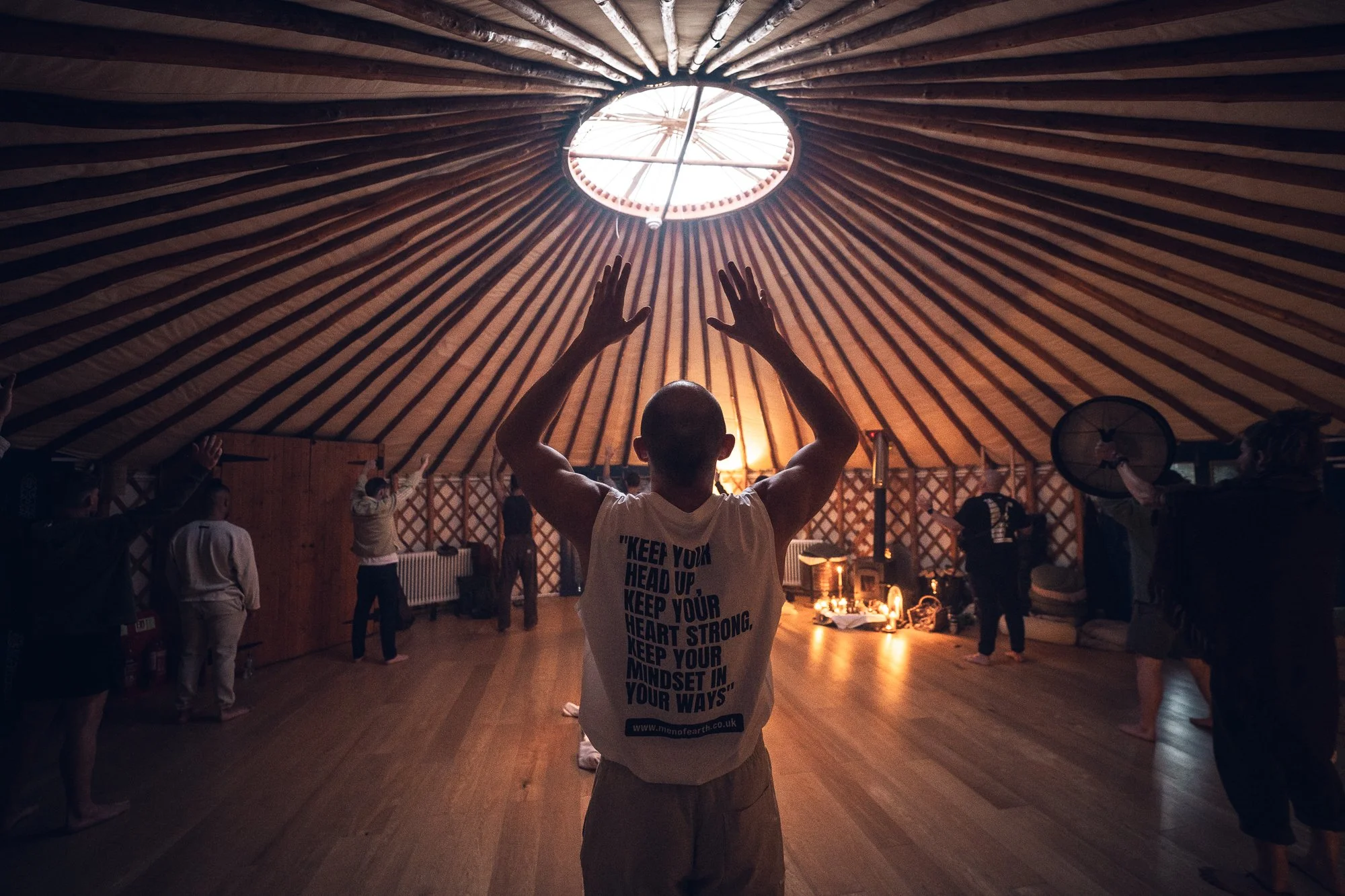 A group of people inside a yurt with a wooden ceiling, participating in a meditation or yoga session. The person in the foreground has their back to the camera, wearing a sleeveless shirt that says 'Keep your head up, keep your heart strong, keep your mindset in your ways.'