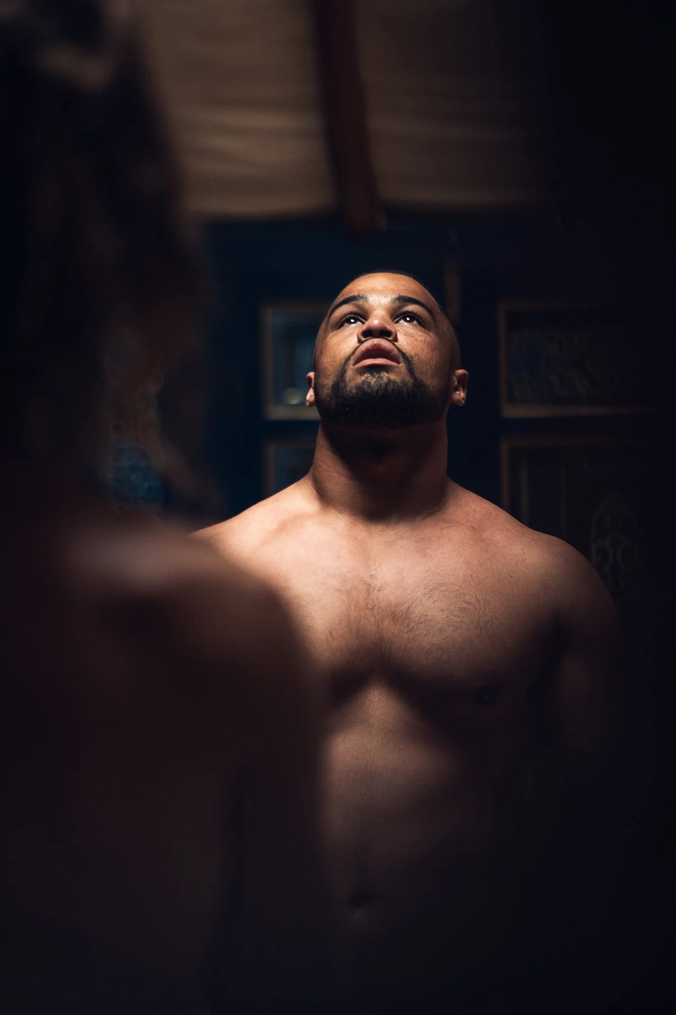 A shirtless man looking upward in a dimly lit room, with blurred foreground of another person.