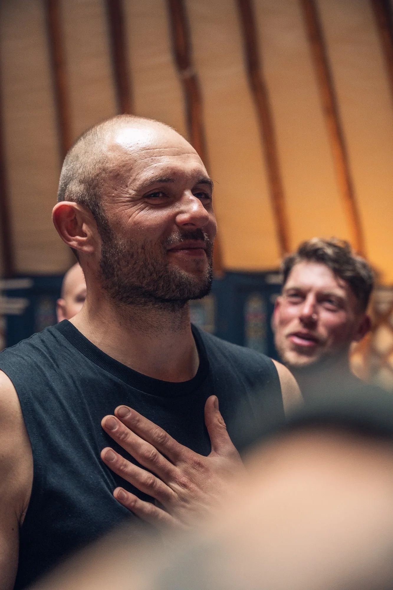 A man with a shaved head and beard holding his hand over his heart, appearing to sing or recite, with a man smiling behind him, in a wooden-paneled room.