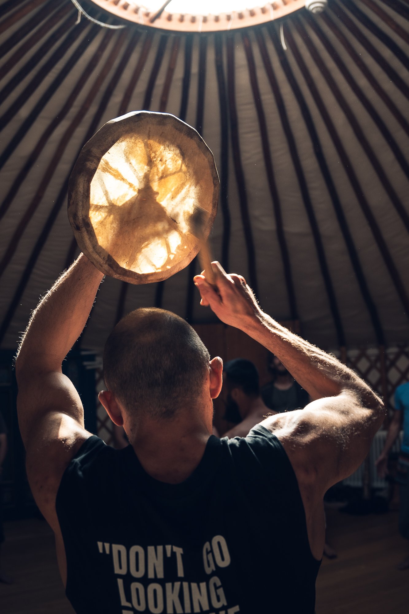 A man playing a drum inside a teepee with a wooden drum, using a drumstick, and people in the background.