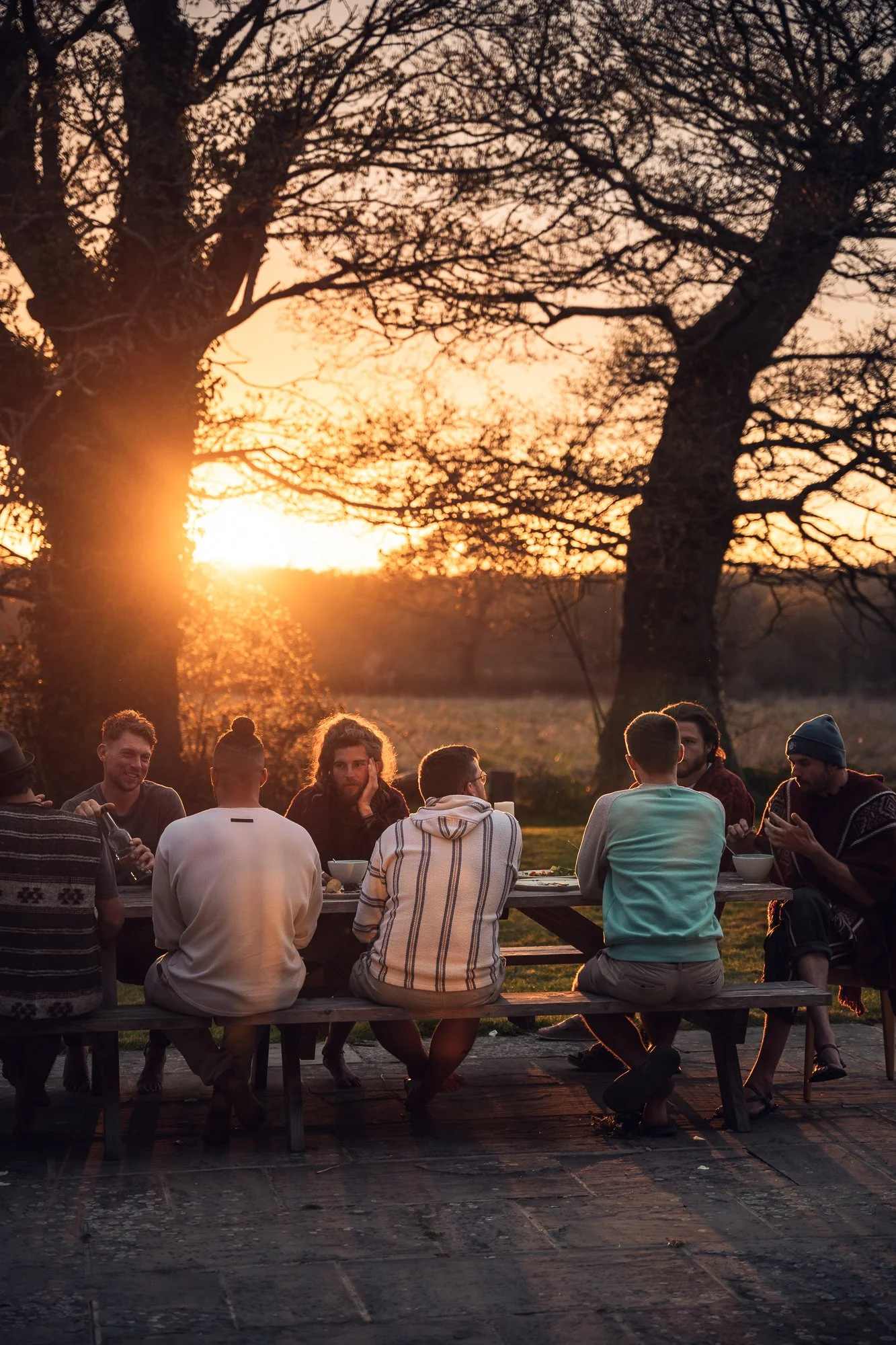 Group of people sitting at a picnic table outdoors during sunset, with bare trees in the background.