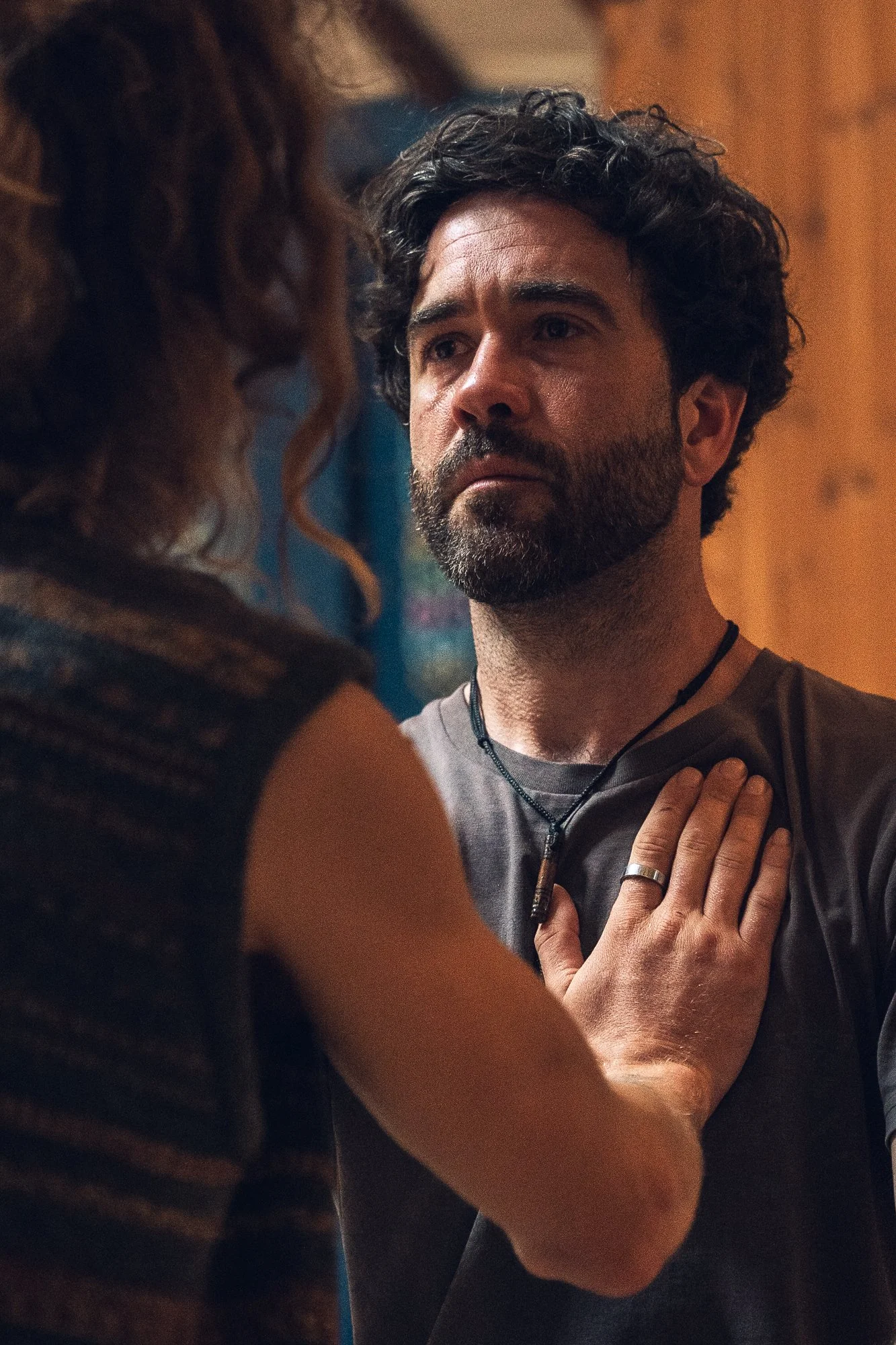 A man with dark curly hair and a beard looks serious as a woman places her hand on his chest.