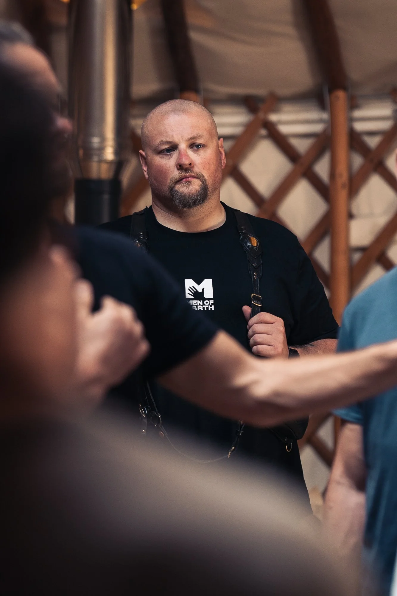 A man with a serious expression, shaved head, and beard, wearing a black T-shirt with a 'Men of Earth' logo, standing among a group of people in what appears to be an indoor gathering or meeting, with wooden lattice and piping in the background.