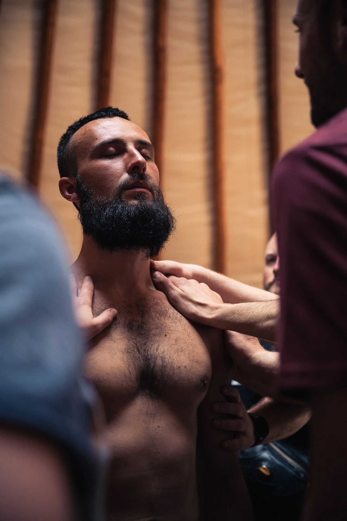 A group of people is touching a shirtless man with a beard, with their hands on his neck and shoulders, in a close-up setting indoors.