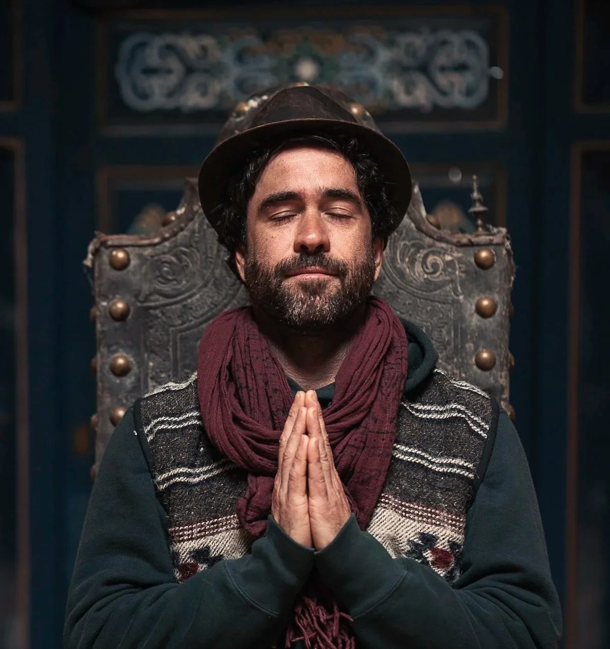 A man with a beard and closed eyes, sitting with hands in prayer position, wearing a hat and maroon scarf, in front of an ornate wooden seat.