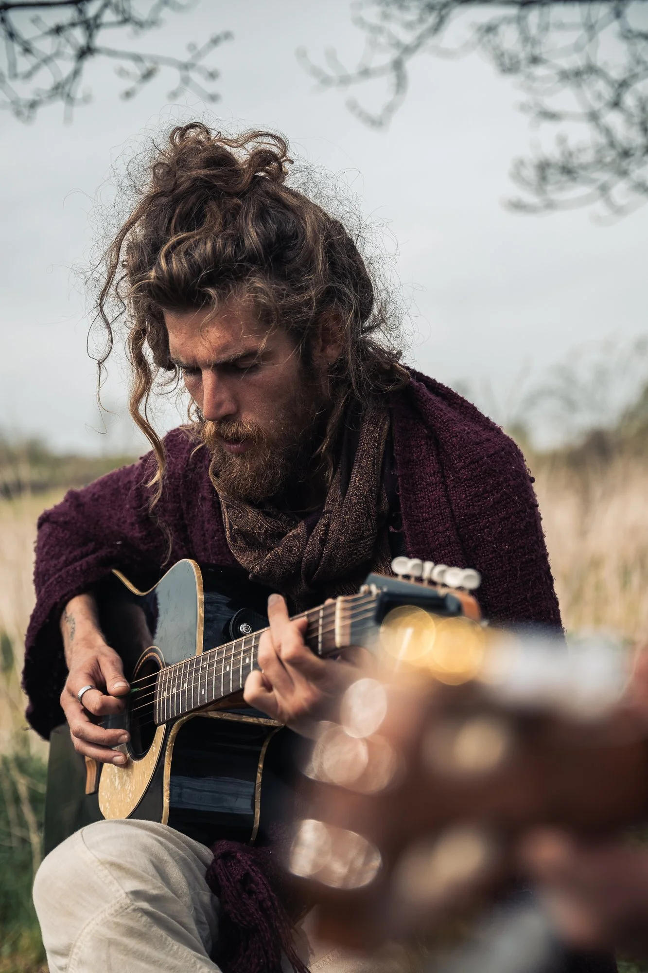 A man with long, curly hair and a beard playing an acoustic guitar outdoors during daytime.