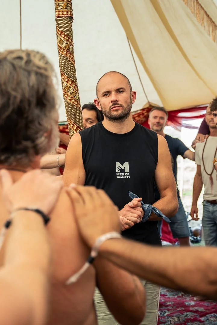 A group of people, mostly men, inside a tent at a gathering. The central figure is a man with a shaved head in a sleeveless black shirt with the logo 'Men of Earth.' Others are around him, some smiling, with a decorative pole and fabric visible in the background.