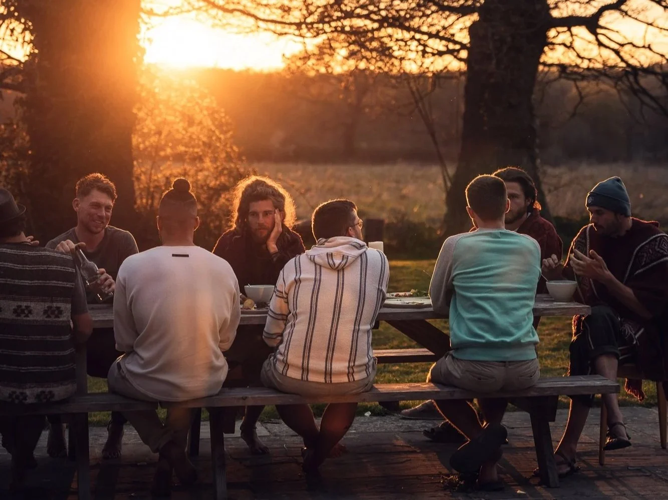 Group of people sitting at a picnic table outdoors during sunset, engaging in conversations and having a meal